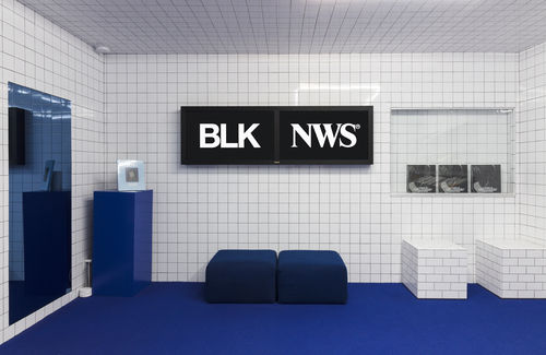 Photograph of room with the walls covered in white, square ceramic tiles. Two square ottoman to the right of the image are also covered in similar tile. The carpet in bottom half of image is navy blue.  There are two small navy blue ottomans side by side. Above them on the tile wall are two video monitors with  the letters BLK on left screen and NWS on right screen. The letters are in white with a black background.
