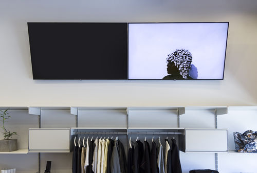 Image of a store counter with clothes on a rack that is framed by a floating, horizontal shelving unit. There a six cubbies, the clothing rack spans the length of two. A plant is on the extreme left cubby. Above the shelving unit are two video monitors.  The left monitor is completely black. The right monitor is a profile view  image of a person with a headdress covered in cowrie shells.