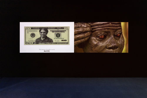 A photograph of two videos projected side-by-side against a black wall that takes up two-thirds of the image. The carpet in the lower third of the photograph is blue.