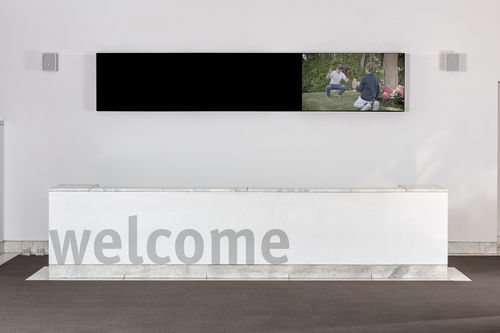 Image of a wall in a large room with white walls and a grey floor. A large white piece of furniture spans nearly the full length of the image and the words "welcome" are printed in grey on front. High on the wall is a long, narrow horizontal black  structure, which appears to be a video screen. On the right 1/3 of this narrow area is an image of a man and a boy playing catch, with baseball gloves, outdoors in a lush, green setting. The man wears jeans and a white T-shirt and squats, and the boy has his back
