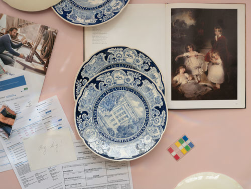 Photograph looking down on a pink surface with objects strewn. At top right is an open book showing an image of a European painting on right and text on left. Two blue-and-white plates sit on top the book in center of the image. At top a portion of another blue-and-white plate is visible. Paper that looks like opened mail and bills is on the left--an index card with "Big Hun!" hand written on it is atop this paper. A colored note flags sits on lower right.
