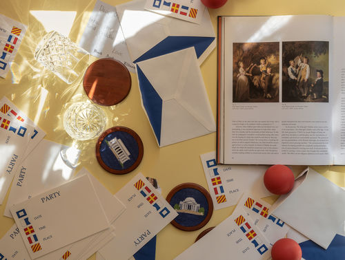 Photo looking down on a yellow surface with objects strewn on it. Several envelopes and cards with the words "PARTY" and nautical-themed graphics are strewn across the surface. At right is a book open to a page showing to images of European paintings with text below.  Several coasters with Washington, D.C. monuments depicted are visible with two etched lowball cocktail glasses. At lower right are two red balls. 