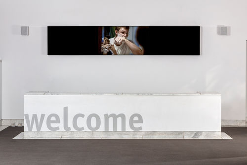 Image of a wall in a large room with white walls and a grey floor. A large white piece of furniture spans nearly the full length of the image and the words "welcome" are printed in grey on front. High on the wall is a long, narrow horizontal black  structure, which appears to be a video screen. On the middle 1/3 of this narrow area is an image of a man and a boy looking at the camera and perhaps in a mirror, wiping a white, foamy material on or off their faces.