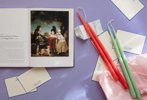 Photo looking down on a purple surface with objects strewn on top. At top left a book is open to a page with a European painting on the right and text on the left.  Index cards, a folded receipt, and pink tissue paper are strewn around, and 2 sets of unburned candles, orange and green, are set on top the tissue paper at right.