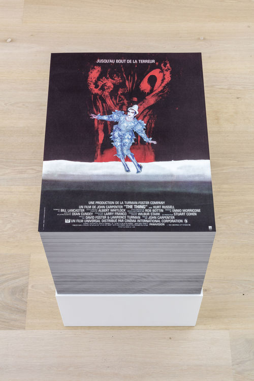 View of a stack of posters sitting on a gallery floor. The posters are a collage of  David Bowie in  the bluish silver clown costume he wore in the "Ashes to Ashes" video and Scary Monsters (and Super Creeps) 1980  album cover. This image of Bowie is on top of the French promotion poster of the 1982 film "La Chose (The Thing)"