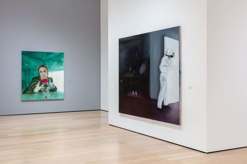 A corner of a gallery, with a green portrait of a woman hanging on a gray wall, and a black-and-white painting hanging on a white wall