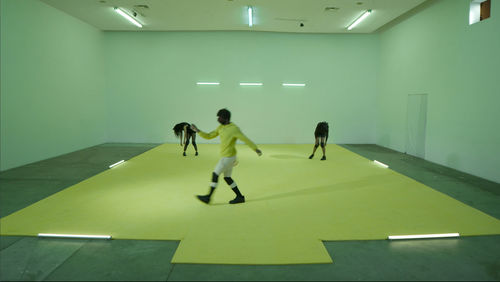 Three dancers with dark skin and dark hair stand in a large room with concrete floors, white walls, a yellow area marked out on the floor, and fluorescent lighting. Two dancers stand in the background, with shoulders and head hunched forward, arms hanging downward, and knees turned inward. A figure in the middle of the foreground is walking across the floor from right to left, wearing a yellow sweatshirt and white shorts.