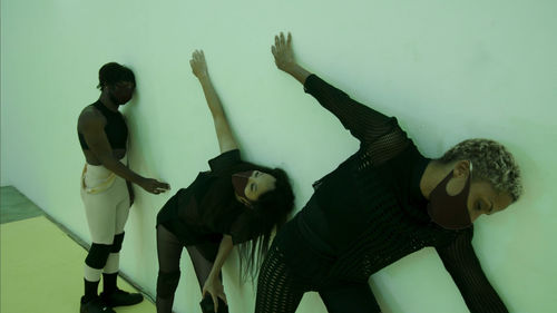 Three figures pose while learning against a white wall. The two foreground figures lean with their backs against the wall, with arms stretched to their sides. The figure in the background stands tall, with with left shoulder  and left side of face against the wall, and the right hand touching the wall.