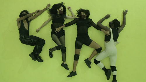 View from above of four figures that lie sprawled on a yellow background. The figures have dark skin and dark hair, wearing black and white clothes and face masks.
