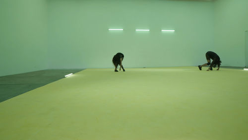 Two dark figures crouch close to the floor in the back of a room with  a yellow floor and three fluorescent lights on the wall in the background.