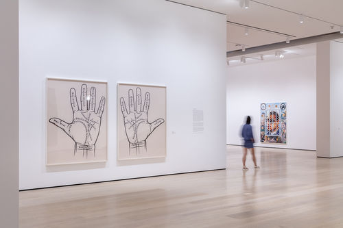 An image inside a white-walled gallery. To the left of the image are two photographs, with a left and right palm in each photo. There are various names written on the palms. In the background is one painting rendered in an airbrush, lowrider car style.