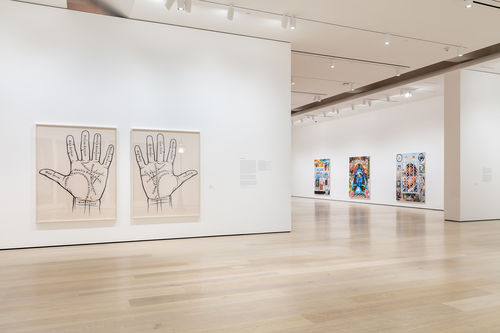 An image inside a white-walled gallery. To the left of the image are two photographs, with a left and right palm in each photo. There are various names written on the palms. In the background are three paintings rendered in an airbrush, lowrider car style.