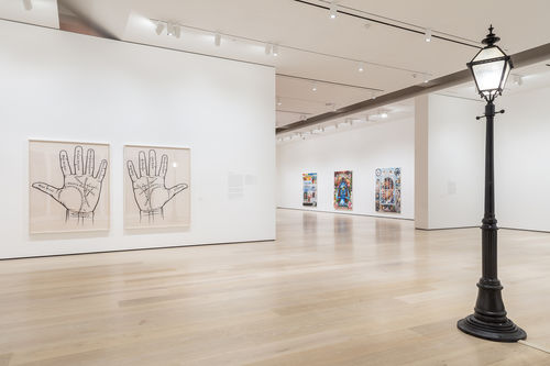 An image inside a white-walled gallery. In the foreground to the right is a black streetlamp. To the left in the middle of the image are two photographs, with a left and right palm in each photo. There are various names written on the palms. In the background are three paintings rendered in an airbrush, lowrider car style.