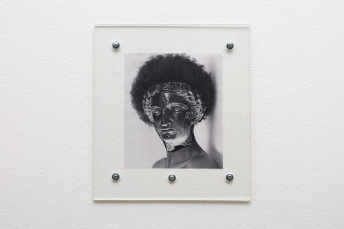 Black and white photograph tacked to a wall under a sheet of clear material depicts a woman with curly afro hair, and the female face of a Greek sculpture  pasted on top of the woman's dace. 