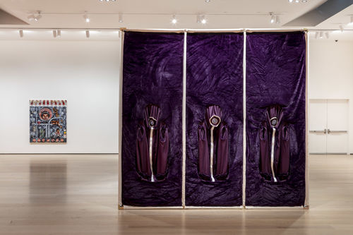 A large gallery with white walls with an artwork on the far wall and in the center of the room a large square structure of white poles and what purple fabric covering the surface. Three panels each have a white circle at head height, with a white stripe down to the floor. 