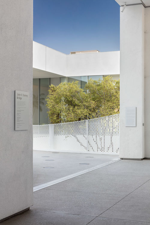 Photo of the Hammer Museum upper gallery terrace, looking towards the bridge, shows white walls and the top of a tree in the background. On the columns on wither side of the bridge entrance there are two large panels of text. 