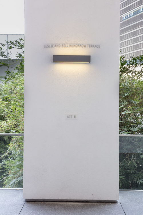 Photo of the Hammer Museum upper gallery terrace, a white architectural column isin the center of the photo with greenery in the background. On the column is the text "ACT II" at about chest height, a light fixture is above head height, and the words "LESLIE AND BILL MORROW TERRACE" above the light. 