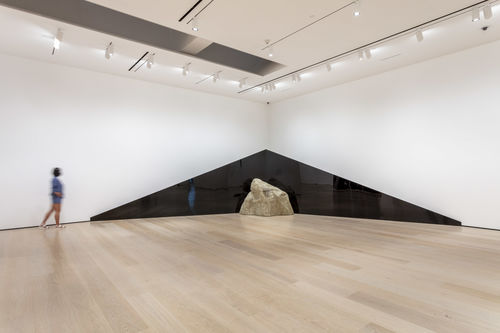 A view of a large gallery, looking into the corner. A dark, triangular shape extends across the corner, with the peak in the corner. A large rock sits on the floor in front of the triangle. A person walks to the left of the work, against the far wall.