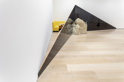 A view of the corner of a gallery. Two dark triangular structures extend across the corner, meeting in the middle at the tall ends. A large rock sits on the floor in front of the triangles, where they meet. Behind the triangles, between them and the wall, a yellow corner booth and a table are visible.