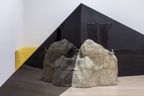 A view of the corner of a gallery. Two dark triangular structures extend across the corner, meeting in the middle at the tall ends. A large rock sits on the floor in front of the triangles, where they meet. A drinking fountain spout extends from the  top of the rock. Behind the triangles, between them and the wall, the edge of a yellow piece of furniture is visible.