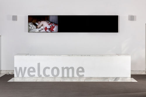 Image of a wall in a large room with white walls and a grey floor. A large white piece of furniture spans nearly the full length of the image and the words "welcome" are printed in grey on front. High on the wall is a long, narrow horizontal black  structure, which appears to be a video screen. On the left 1/3 of this narrow area is an image of a man lyng on his stomach in bed, face in a pillow, lying under a red and white quilt. At left and right of the black shape are grey cubes that appear to be speakers