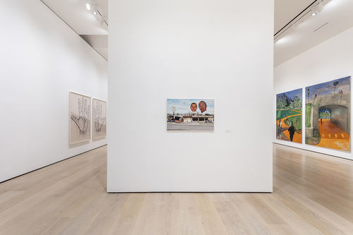 A photograph of the inside of a white-walled gallery where a vertical wall splits the gallery  (and image) in two. On the wall is a painting. On either side of the wall when one rounds the corner are artworks.