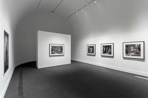 A white gallery with a curved back wall with three black and white photographs hanging on the far wall, one hanging on the wall to the left, and one on a free-standing wall near the back of the room. 