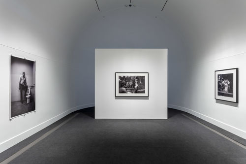 A white gallery with a curved back wall with one black and white photographs hanging on the wall at right, one hanging on the wall to the left, and one on a free-standing wall near the back of the room. 