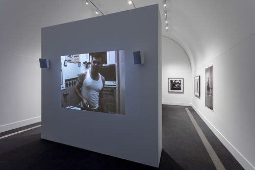A white gallery with a free-standing wall standing in the foreground. A projection of a color image is visible on the free-standing wall. Three black and white photographs are visible on the gallery walls to the right in the background.