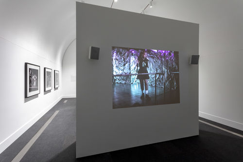 A white gallery with a free-standing wall standing in the foreground. A projection of a color image is visible on the free-standing wall. Three black and white photographs are visible on the gallery walls to the left in the background.