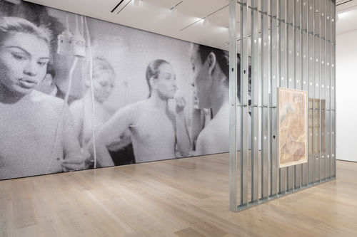 A large gallery with a black and white floor-to-ceiling photomural on the back wall. In the mural, four people are visible from the waist up with hair tied back. One is applying makeup. A power cord hangs across the foreground on the left. In the foreground of the gallery is a wall of metal studs with an arwork of muted colors hanging on it.