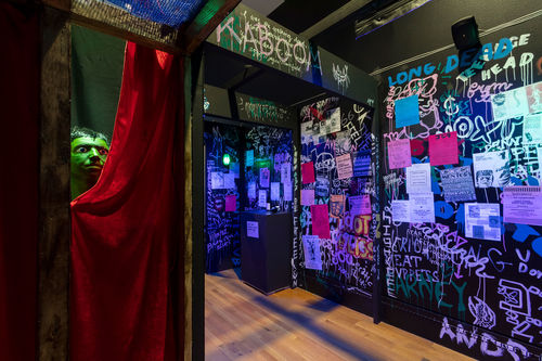 A green head pokes out from behind a red curtain in a flyer- and graffiti-covered haunted house