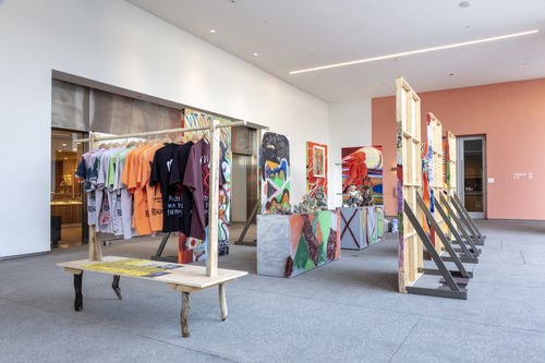 Photo of a an outdoor terrace with an installation of temporary wooden walls, a coatrack with t-shirts hanging on it and colorfully painted pedestals with objects on top.