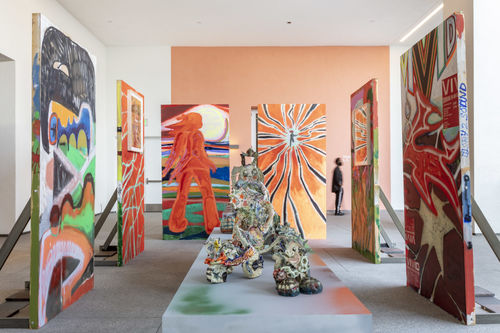 Photo of a an outdoor terrace with an installation of temporary wooden walls, a coatrack with t-shirts hanging on it and colorfully painted pedestals with objects on top.