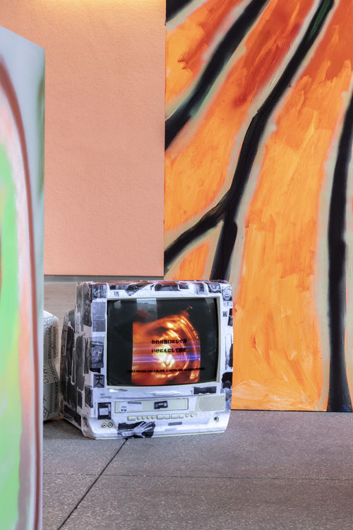 Photo of a TV set sitting on a concrete floor, with black and white images pasted on the body and an orange image on the screen. To the right of the TV is an orange and black painted surface, and a pale orange wall is in the background.