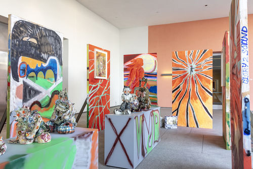 Photo of a an outdoor terrace with an installation of temporary wooden walls, a coatrack with t-shirts hanging on it and colorfully painted pedestals with objects on top.