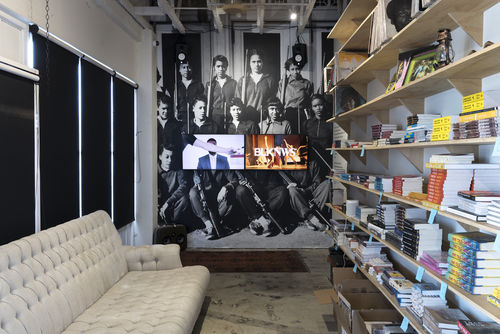 Image of a narrow, shotgun room with a wooden bookshelf on the right populated with books and creamy, white couch on the left. At the backwall is a wallpaper of a black and white photograph of women with rifles. There are two television screens in the middle of the wall with news footage.