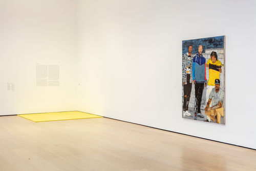 A yellow pad on the floor in the corner of a gallery, with a colorful portrait hanging nearby