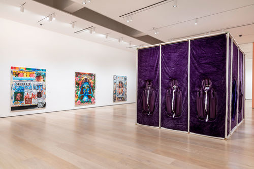 A large purple cube in a gallery with three colorful artworks hanging on the wall
