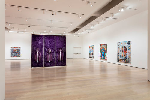 A large purple cube in a gallery with four colorful works hanging on the wall