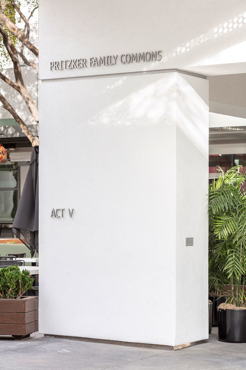 Photo of Hammer Courtyard with a white column in the center of image. In middle of column the words "ACT V" appear in grey three-dimensional letters. At top of the column are the words "PRITZKER FAMILY COMMONS" in the same grey letters.