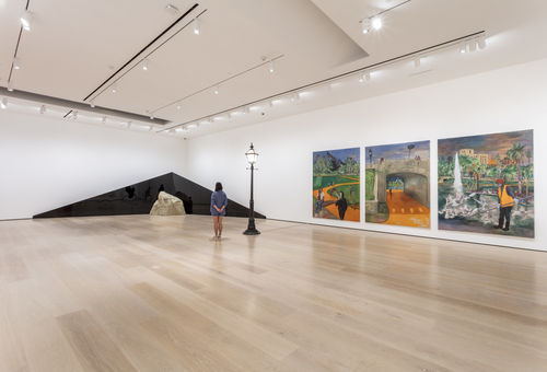 A gallery with a black streetlamp in the middle that divides two works. To the right is a triptych of paintings displaying MacArthur Park in Los Angeles. To the left is a dark, triangular shape that extends across corner of the room, with the peak of the object in the corner/center. A large rock sits on the floor in front of the triangle. A person stands to the left of the streetlamp.