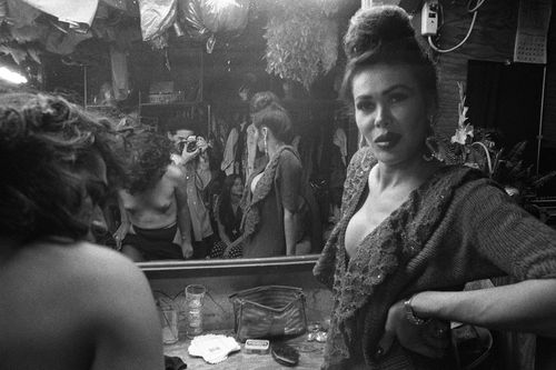 A woman in makeup looks over her shoulder with a mirror behind her, the photographer visible in the mirror