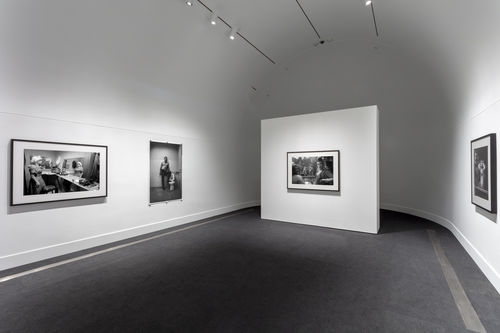 A white gallery with a curved back wall with one black and white photograph hanging on the central, freestanding wall, two black and white photographs on the left wall, and one photograph on the right wall.