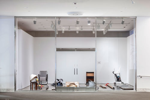 Photograph of a gallery with a glass wall, viewed from the outside. Various objects. are sitting on the floor of the gallery.
