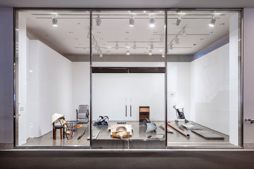 View of a gallery with a glass wall from the outside. Various objects  are sitting on the floor of the gallery.