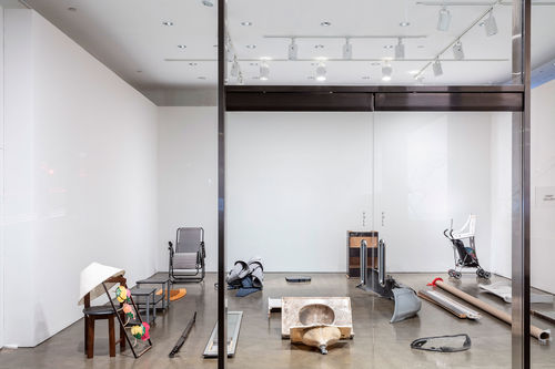 View looking into a room through a glass wall and glass doors. The room has various objects sitting on the floor, such as a lamp, a sink, a chair, blankets.