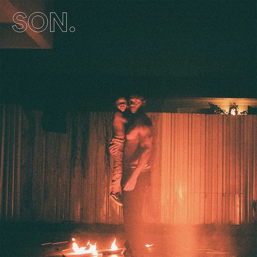 Dark photograph outside, at nighttime, of a man holding a small child, while standing in front of a fire in the foreground. The man is looking at the viewer, and the fire reflects against a fence in the background. Words are on the image: "SON." in the top left corner,