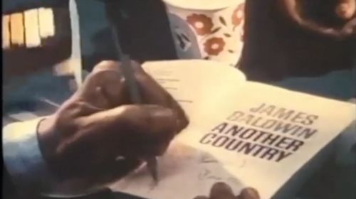 Image of a person's left hand signing the inside cover page of James Baldwin's book, Another Country. The book rests on a table with a dark blue tablecloth. A white paper cup with flowers is to the top left of the book.