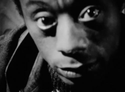 A close crop black and white portrait of James Baldwin. His eyes and eyebrows frame the top of the image and his chin the bottom. He stares directly at the camera. To the left of his face are articles of clothing--perhaps a dress shirt, sweater, and jacket--that are layered.